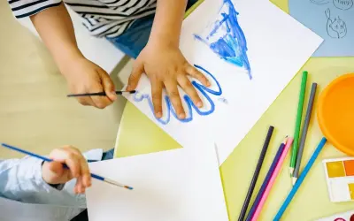 Manos de niños pintando, creando sus propios dibujos para imprimir. Lápices de colores y acuarelas a la vista.