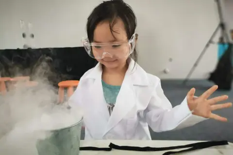 Niña con bata y gafas realiza experimentos para niños de 7 años, creando humo blanco de un cubo.