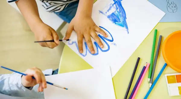Manos de niños pintando, creando sus propios dibujos para imprimir. Lápices de colores y acuarelas a la vista.
