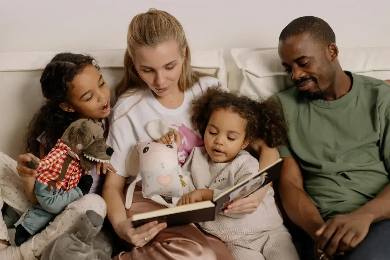 Familia reunida leyendo cuentos para niños de 8 años. La madre lee un libro a sus dos hijas y al padre, rodeados de peluches.