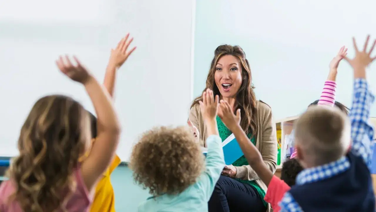 Maestra animada interact&uacute;a con ni&ntilde;os que levantan las manos, participando en divertidos juegos de lengua en el aula.