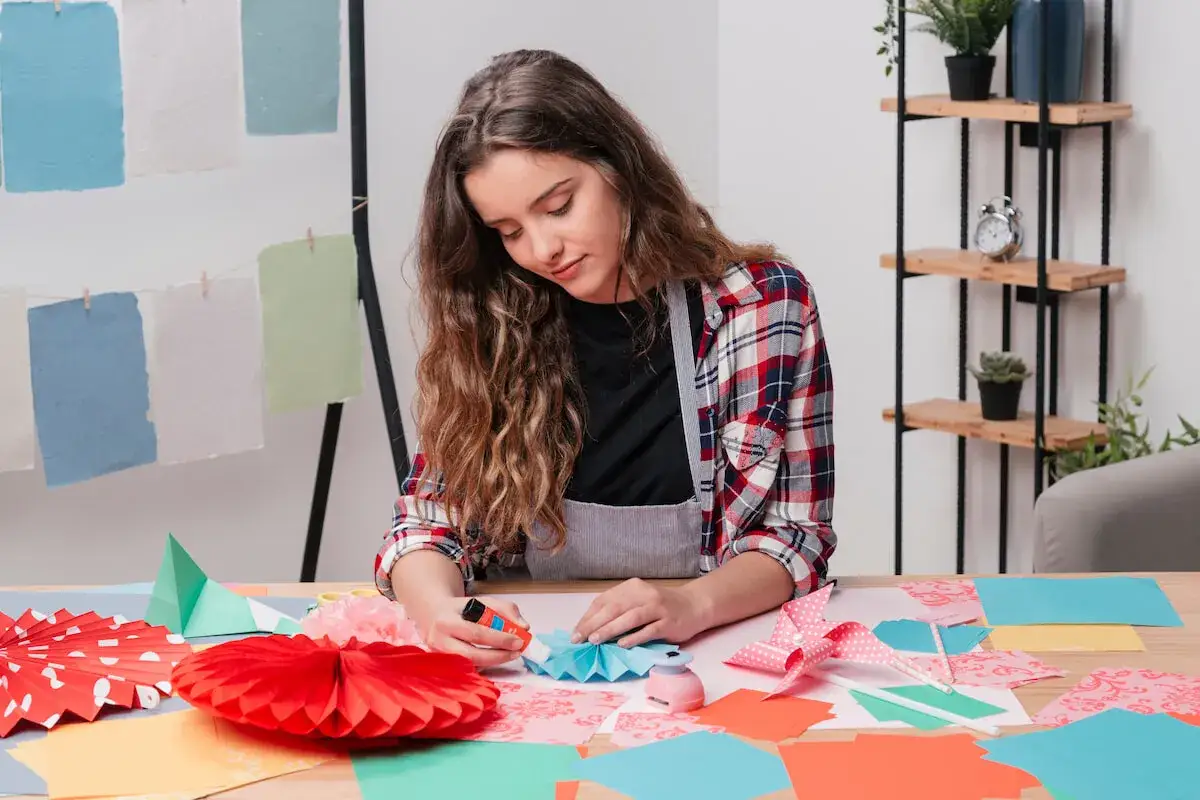 Chica creando manualidades para ni&ntilde;os de 12 a 15 a&ntilde;os. Papeles de colores, abanicos y pegamento sobre la mesa.