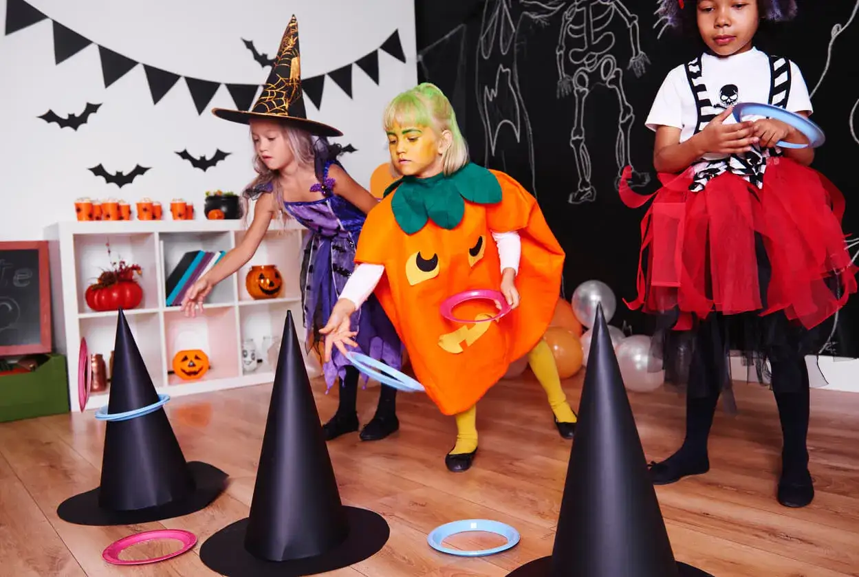 Ni&ntilde;as disfrazadas de bruja y calabaza juegan a lanzar aros en sombreros de bruja, parte de divertidos juegos de Halloween para ni&ntilde;os.