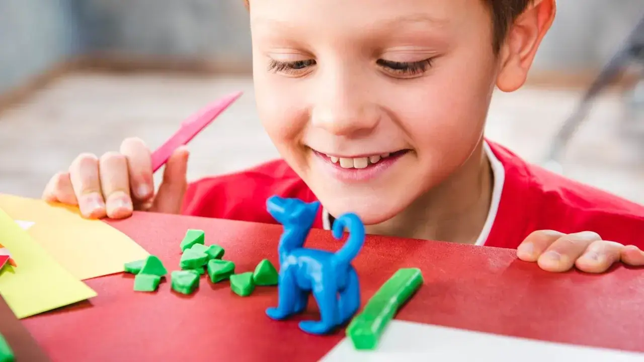 Niño sonriente crea un dinosaurio azul con plastilina casera, rodeado de piezas verdes y herramientas de manualidades.